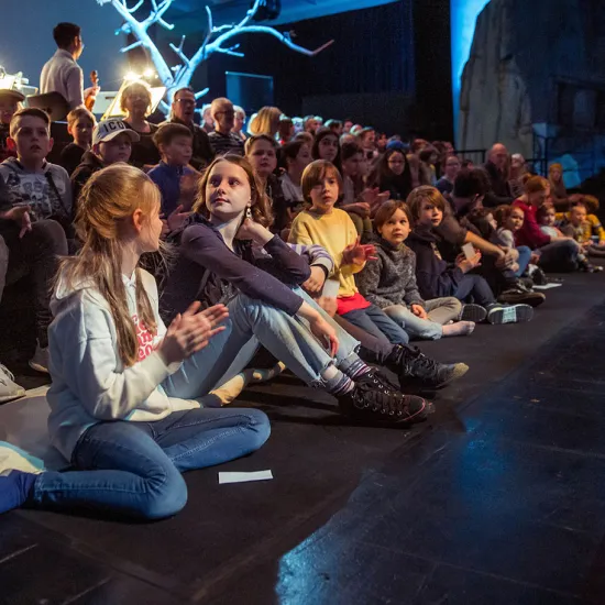 Several children are sitting on the floor directly in front of the stage, watching the performance attentively. Behind them, more children and adults are seated on tiered rows, following the performance. In the background, parts of the orchestra and a leafless stage tree are visible. The scene conveys the focused atmosphere of a family performance.