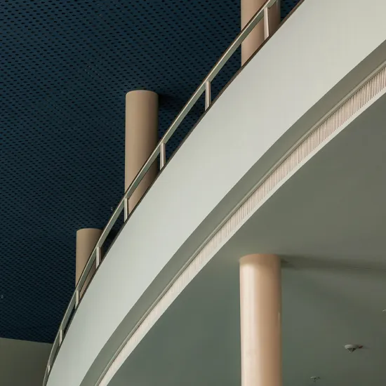 View of the foyer of the Cologne Opera at Offenbachplatz. Curved gallery levels with light-coloured balustrades and slender columns rise beneath a dark blue perforated ceiling. The architectural details emphasise the clean lines and modern design of the building.