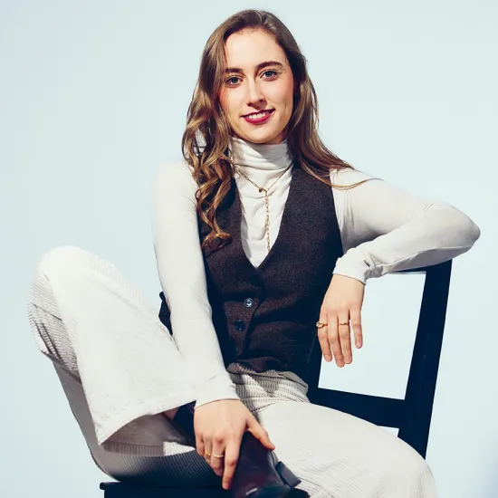 Portrait of soprano Lana Sophie Westendorf seated on a black chair against a light grey background. She is wearing a white turtleneck, a dark waistcoat and light-coloured trousers, with one arm resting casually on the back of the chair. She looks directly at the camera with a warm smile, creating a relaxed and approachable impression.