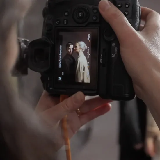 Photographer Teresa Rothwangl holds a camera in her hands and looks at the image displayed on its screen. Visible is a portrait of two performers, taken during the photoshoot for the campaign visual of Der Rosenkavalier, one of Opera Cologne's premiere productions for the 2026/27 season.