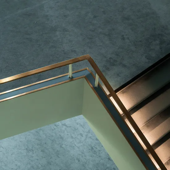 View of the stairwell at the Cologne Opera at Offenbachplatz. A light-coloured gallery with a brass handrail forms a striking angle beneath a dark textured ceiling. An illuminated staircase runs alongside it, highlighting the building's clean, modern architecture.
