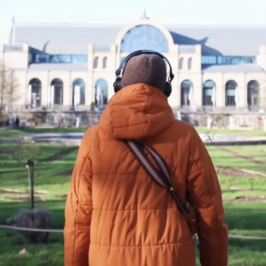 oprano Alina König Rannenberg stands with headphones on and her back to the camera in the gardens of Cologne’s Flora. The Festhaus can be seen in the background.