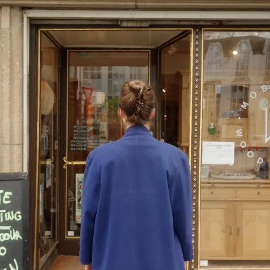 Opera singer Johanna Thomsen, seen from behind wearing a blue coat, stands outside the antiques and furniture restoration shop Modanese Wood and Wine. Restored wooden furniture is displayed in the shop window.