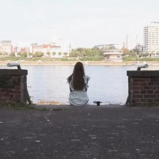 Mezzo-soprano Tina Drole sits with her back to the camera on the banks of the Rhine in Deutz’s Rheinpark, looking across the river towards Cologne’s city centre.