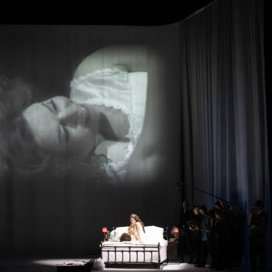 Auf der Bühne sitzen die Feldmarschallin Fürstin Werdenberg (Magdalena Hinterdobler) und Octavian (Bianca Andrew) in einem Bett. Über ihnen ist eine Live-Schwarz-Weiß-Nahaufnahme der Feldmarschallin projiziert. Szene aus Richard Strauss’ „Der Rosenkavalier“.