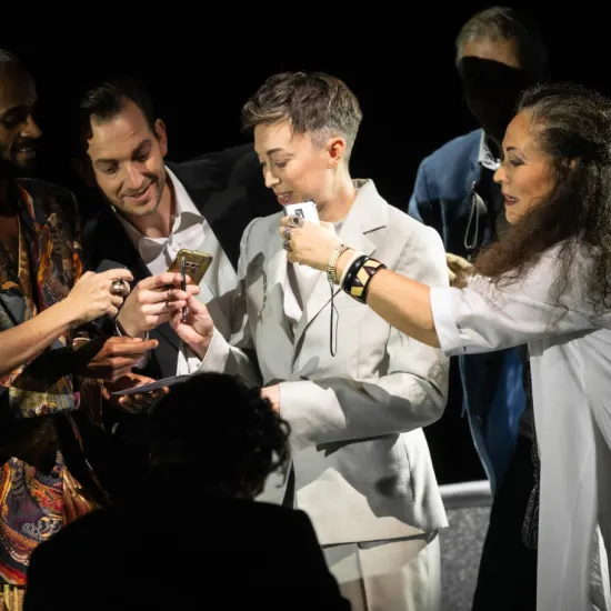 Octavian (Bianca Andrew) steht im hellen Anzug inmitten der Statisterie der Oper Köln. Mehrere Personen halten ihr Gegenstände entgegen; Annina (KS Dalia Schaechter) steht rechts neben ihr. Szene aus Richard Strauss’ „Der Rosenkavalier“.