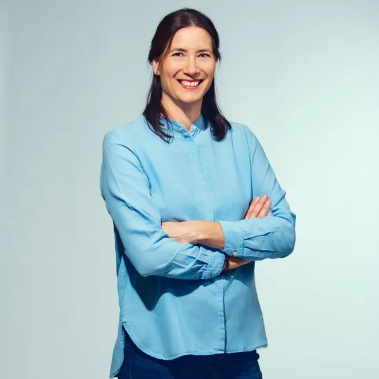 Portrait of Brigitta Gillessen against a light grey background. She is wearing a light blue blouse and dark blue trousers, standing with her arms crossed and smiling at the camera.
