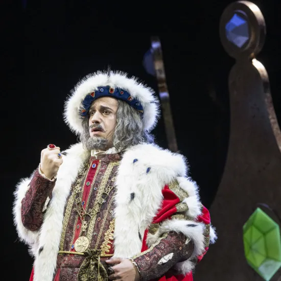 The old King Detlef (Ferhat Baday) raises his ring-adorned fist. He wears a red and gold robe trimmed with white fur, a crown, and a grey wig with a beard. Scene from the children’s opera “DER KÖNIG VERSCHENKT WAS” by Philipp Matthias Kaufmann.