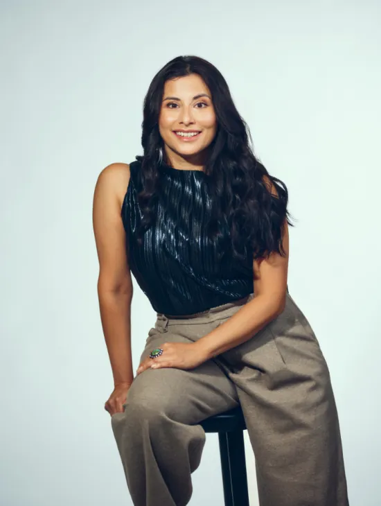 Mezzo-soprano Adriana Bastidas-Gamboa is seated on a black stool against a light grey studio background. She looks directly at the camera with a warm smile. Her long, dark, wavy hair falls over her shoulders. She wears a dark, subtly shimmering sleeveless top and wide-legged grey trousers. A large ring and hoop earrings stand out as distinctive accessories. The portrait conveys an open, confident, and approachable impression.