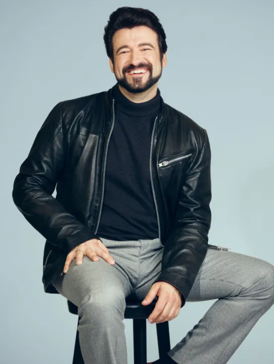 Tenor Dmitry Ivanchey is seated on a stool, smiling warmly at the camera. He is wearing a dark turtleneck sweater, a black leather jacket, and light grey trousers. His dark hair and neatly groomed beard give the portrait a distinctive appearance. Set against a light blue background, the photograph has a friendly, modern, and professional look.