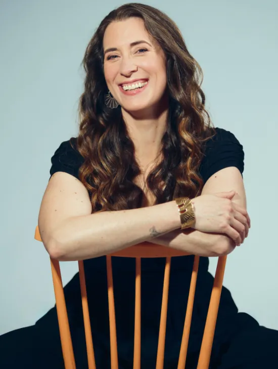 Soprano Emily Hindrichs sits behind a light wooden chair, resting her arms on the backrest. She smiles warmly at the camera. Her long, wavy brown hair falls over her shoulders. She is wearing a dark dress, distinctive earrings, and a wide bracelet. Set against a light blue background, the portrait has a friendly, relaxed, and professional appearance.