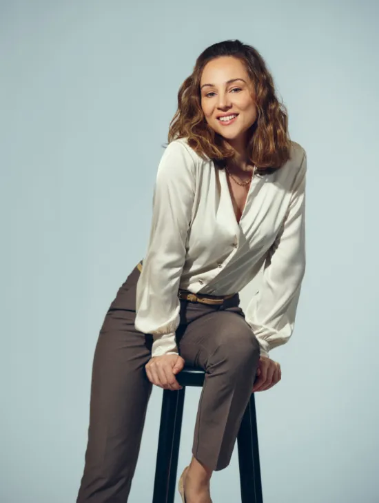 Soprano Giulia Montanari sits relaxed on a black stool against a light grey studio background. She leans slightly forward, smiles at the camera, and wears a cream-coloured, subtly lustrous long-sleeved blouse paired with grey trousers. Her shoulder-length, softly wavy hair frames her face naturally. The portrait conveys an open and friendly impression.