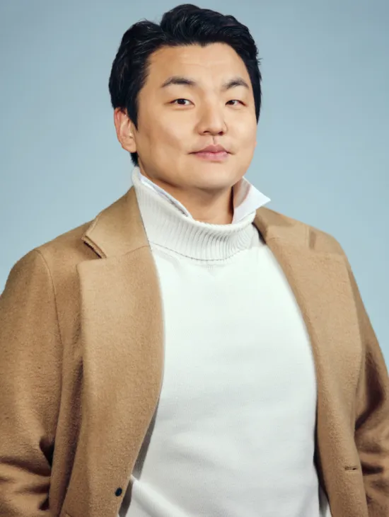Baritone Insik Choi looks directly into the camera. He is wearing a light-coloured turtleneck sweater beneath a beige coat. His dark hair is combed back. Set against a light blue background, the portrait has a calm and professional appearance. The photograph shows him from the upper body upwards.