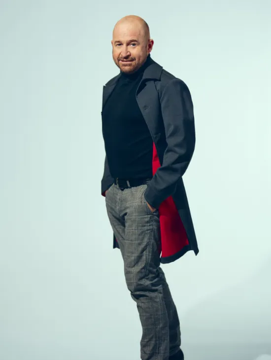 Tenor John Heuzenroeder stands slightly turned towards the camera, looking at the viewer with a subtle smile. He is wearing a dark turtleneck sweater, grey checked trousers, and a dark coat with a red lining. His hands are tucked into his trouser pockets. Set against a light blue background, the portrait has a modern and professional appearance. The photograph shows him in an almost full-length pose.
