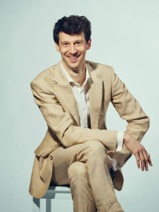Baritone Wolfgang Stefan Schwaiger is seated on a light-coloured stool against a light grey studio background. He looks directly into the camera with a smile and wears a light beige suit with a white shirt. His legs are crossed, and his arms rest casually in front of him. His dark, slightly wavy hair and distinctive moustache are defining features of his appearance. The portrait conveys an open, friendly, and confident impression.