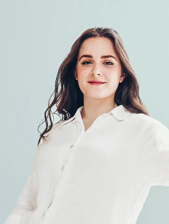 Porträt der amerikanischen Mezzosopranistin Anna Kelly. Sie blickt mit einem leichten Lächeln direkt in die Kamera und trägt eine weiße Bluse. Vor einem hellen, einfarbigen Hintergrund fällt ihr langes dunkles Haar locker über die Schultern.