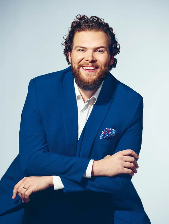 Portrait of bass Christoph Seidl against a light grey studio background. He is seated, leaning slightly forward, smiling directly at the camera, and wearing a bright blue suit with a white shirt and a patterned pocket square. His curly brown hair and full beard are defining features of his appearance. His relaxed posture conveys openness and confidence.
