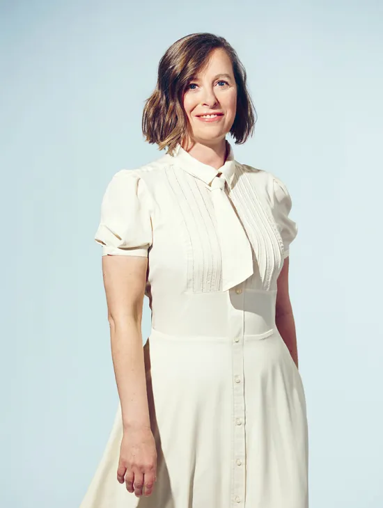 Portrait of soprano Claudia Rohrbach against a light grey studio background. She is standing at a slight angle, looking directly into the camera with a warm smile. She is wearing a cream-white dress with short puff sleeves, fine pintucks, and a bow at the collar. Her chin-length, slightly wavy brown hair is parted to one side. Her upright posture conveys elegance and composure.