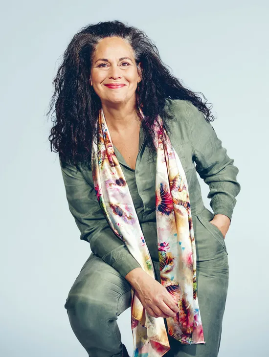 Portrait of Dalia Schaechter. The mezzo-soprano smiles at the camera while sitting slightly leaning forward. She wears an olive-green trouser suit with a long, colourful patterned scarf. Her long, curly dark hair falls loosely over her shoulders. The background is light blue and uncluttered.