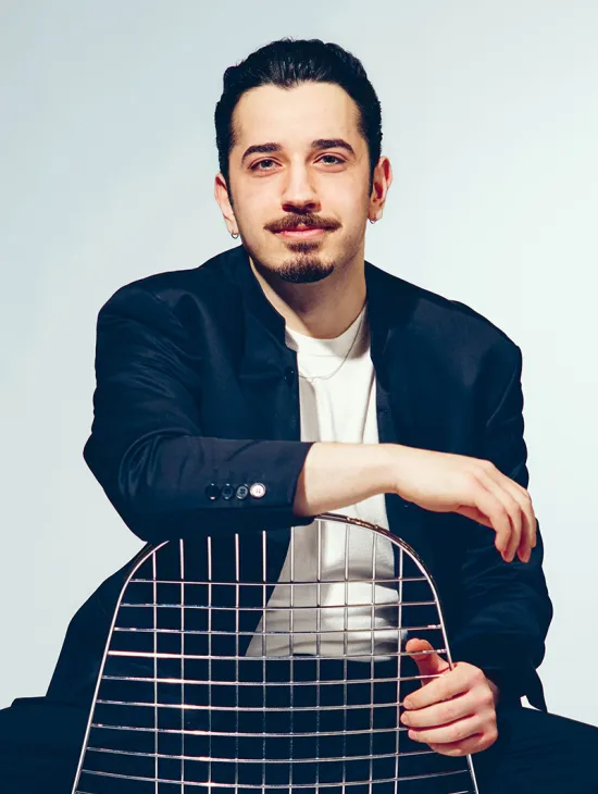 Porträt des Bassisten Ferhat Baday vor hellgrauem Studiohintergrund. Er sitzt hinter der Rückenlehne eines Metallstuhls, auf der er beide Arme locker ablegt, und blickt mit einem dezenten Lächeln direkt in die Kamera. Er trägt ein dunkles Sakko über einem weißen T-Shirt. Sein dunkles Haar ist nach hinten frisiert, dazu trägt er einen Schnurrbart und einen Kinnbart sowie kleine Ohrringe. Die entspannte Haltung vermittelt Ruhe und Selbstbewusstsein.