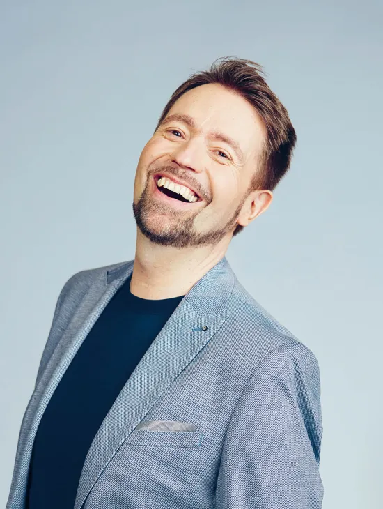 Portrait of Kammersänger Miljenko Turk. The baritone smiles with his mouth open and looks slightly upward. He wears a light blue blazer over a dark T-shirt. His short brown hair is styled back, and he has a neatly trimmed beard. The background is light blue and uncluttered.