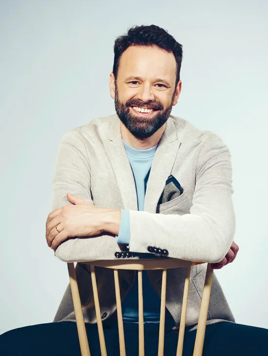 Portrait of Lucas Singer. The bass smiles directly at the camera while resting his folded arms on the back of a light wooden chair. He wears a light grey blazer over a light blue shirt. His dark hair is cut short, and he has a neatly trimmed full beard. The background is light blue and uncluttered.