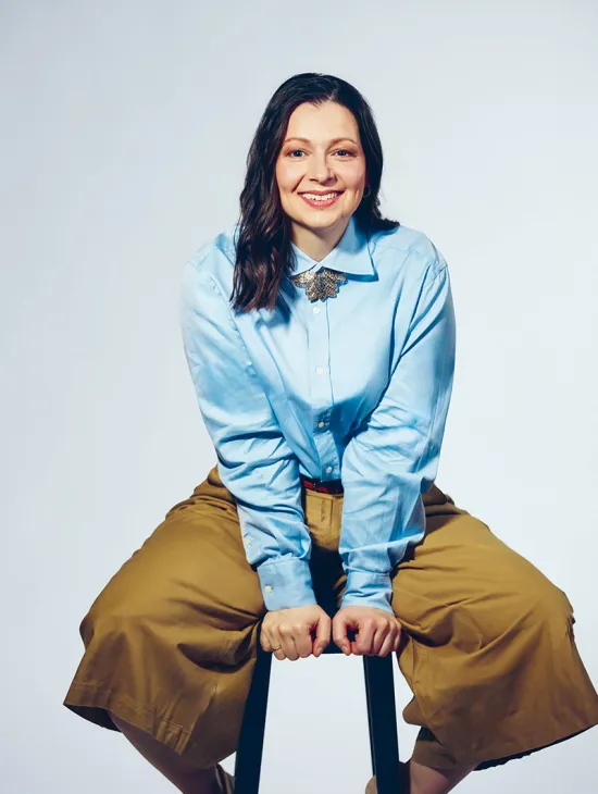 Portrait of soprano Maike Raschke against a light grey studio background. She is seated on a stool, leaning slightly forward and smiling directly at the camera. She is wearing a light blue blouse with a distinctive brooch at the collar and wide beige trousers. Her shoulder-length dark hair falls loosely over her shoulders. Her open posture conveys friendliness and natural ease.