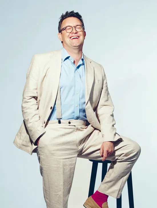 Portrait of Martin Koch. The tenor smiles with his mouth open and looks slightly upward. He wears a light-coloured linen suit with a light blue shirt, sits casually on a black stool, and has one hand in his trouser pocket. His bright pink socks stand out. The background is light blue and uncluttered.