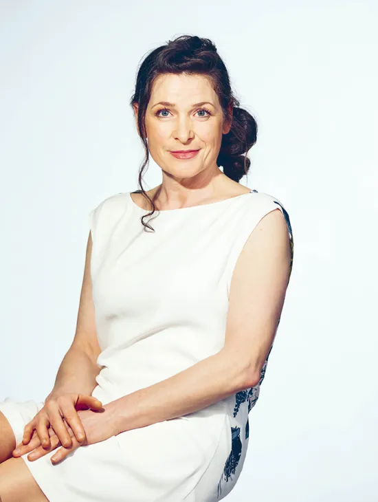 Portrait of Regina Richter. The mezzo-soprano sits with her hands loosely folded and looks into the camera with a gentle smile. She wears a sleeveless white dress with a subtly patterned back panel. Her dark hair is styled in a loose bun. The background is light blue and uncluttered.