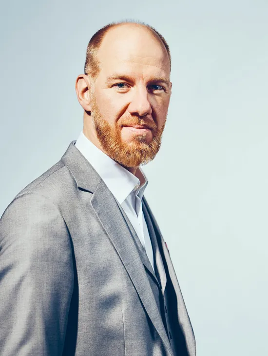 Portrait of Tijl Faveyts. The bass looks directly at the camera with a subtle smile. He wears a light grey blazer over a white shirt and a grey waistcoat. His reddish-blond beard is neatly trimmed, and his hair is cut very short. The background is light blue and uncluttered.