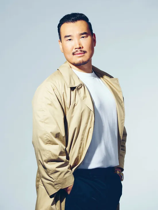 Portrait of tenor Young Woo Kim against a light grey studio background. He is standing at a slight angle to the camera, looking directly into the lens with a calm expression. He is wearing a beige coat over a white T-shirt and dark trousers. His short black hair, moustache, and goatee are defining features of his appearance. His relaxed posture, with one hand in his trouser pocket, conveys composure and confidence.
