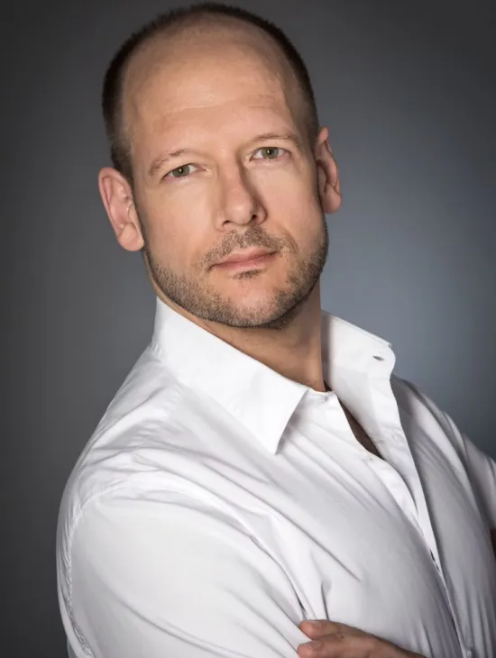 Ein Porträtfoto von Bariton Christian Miedl. Er blickt direkt in die Kamera, trägt ein weißes Hemd mit offenem Kragen und hat kurze Haare und einen Stoppelbart.