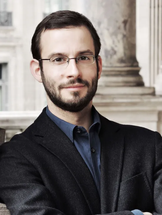 Porträt von Felix Bender. Er trägt eine schmale, rechteckige Brille, einen gepflegten Vollbart und ein dunkles Sakko über einem blauen Hemd. Er blickt ruhig in die Kamera, während im unscharfen Hintergrund klassische Gebäudearchitektur mit einer Säule zu sehen ist.