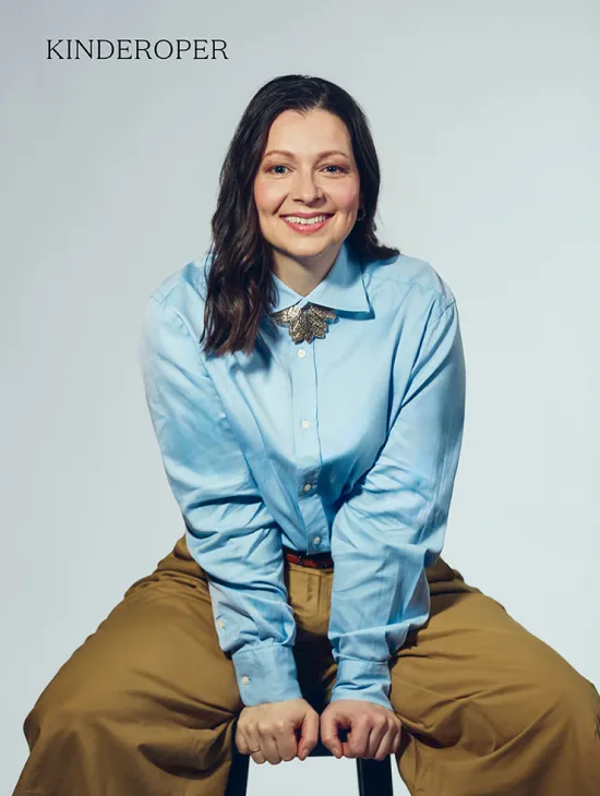 Portrait of soprano Maike Raschke against a light grey studio background. She is seated on a stool, leaning slightly forward and smiling directly at the camera. She is wearing a light blue blouse with a distinctive brooch at the collar and wide beige trousers. Her shoulder-length dark hair falls loosely over her shoulders. Her open posture conveys friendliness and natural ease.