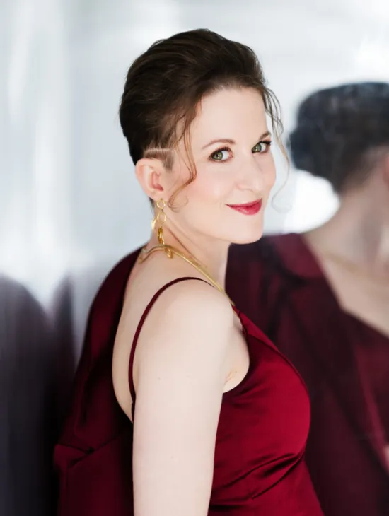 Porträt von Alina Wunderlin in einem roten Kleid. Sie blickt lächelnd über ihre Schulter in die Kamera; im Hintergrund ist ihr Spiegelbild zu sehen.