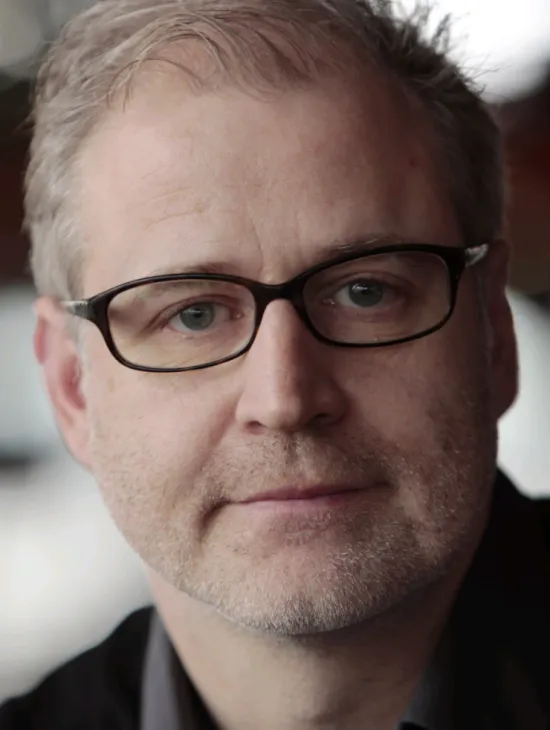 Nahaufnahme von Andreas Grüter mit kurzem grauem Haar, Bartstoppeln und schwarzer Brille. Er blickt vor einem unscharfen hellen Hintergrund in die Kamera.