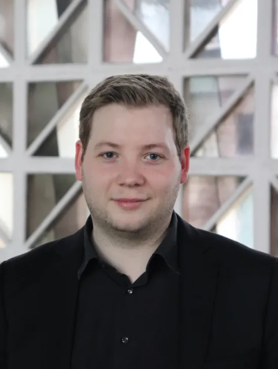 Portrait of Benedict Nagel wearing a black shirt and black jacket, against a bright background with geometric structures.