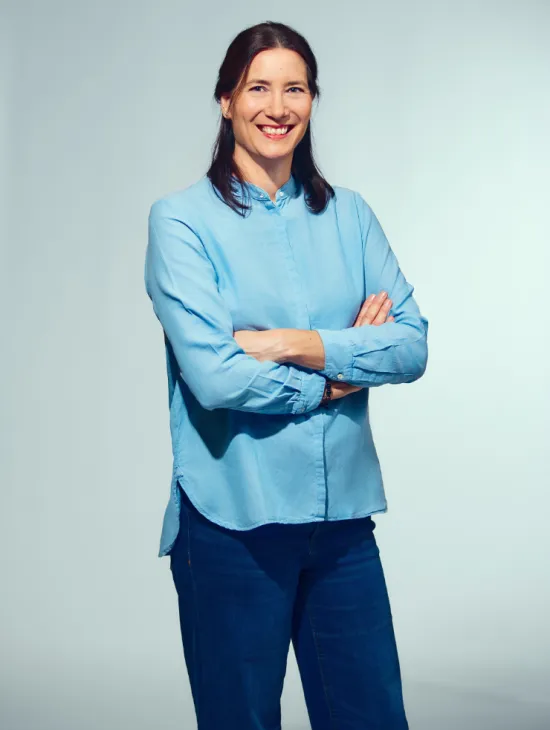 Porträt von Brigitta Gillessen vor einem hellgrauen Hintergrund. Sie trägt eine hellblaue Bluse und eine dunkelblaue Hose, steht mit verschränkten Armen und lächelt in die Kamera.