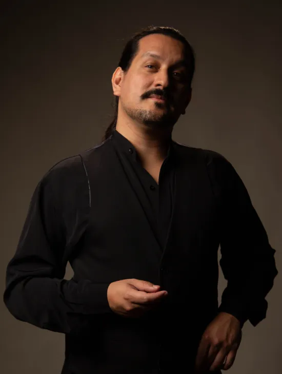 Porträt des Baritons Germán Enrique Alcántara vor einem dunkelbraunen Hintergrund. Er trägt ein schwarzes Hemd mit schwarzer Weste und blickt mit leicht erhobenem Kopf in die Kamera. Sein dunkles Haar ist zurückgebunden, zudem trägt er einen Schnurr- und Kinnbart.