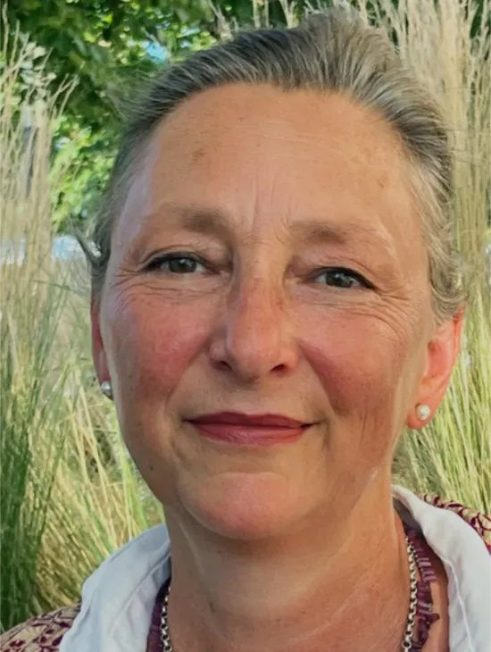 Close-up portrait of Judith Peter with her grey hair tied back and a slight smile. Grasses and green leaves can be seen in the background.