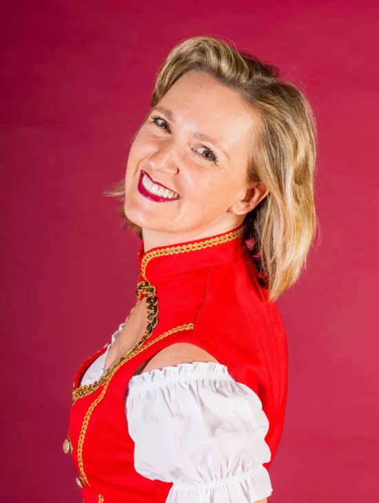 Portrait of Katrin Bachmann wearing a red-and-white costume with gold trim. She looks over her shoulder and smiles at the camera against a pink background.
