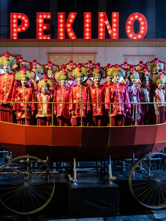 Knaben und Mädchen der Kölner Dommusik in roten Kostümen mit goldfarbenem Kopfschmuck auf einem Bühnenwagen. Im Hintergrund leuchtet der Schriftzug „PEKINO“.