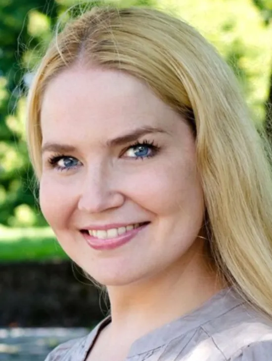 Porträt der Sopranistin Kristi Anna Isene mit langen blonden Haaren und grauer Bluse. Sie lächelt in die Kamera; im Hintergrund ist unscharfes Grün zu sehen.