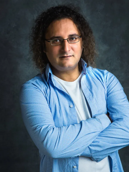 Portrait of Rustam Samedov against a dark grey background. He is wearing glasses and a blue-and-white checked shirt over a white T-shirt, looking at the camera with his arms crossed.