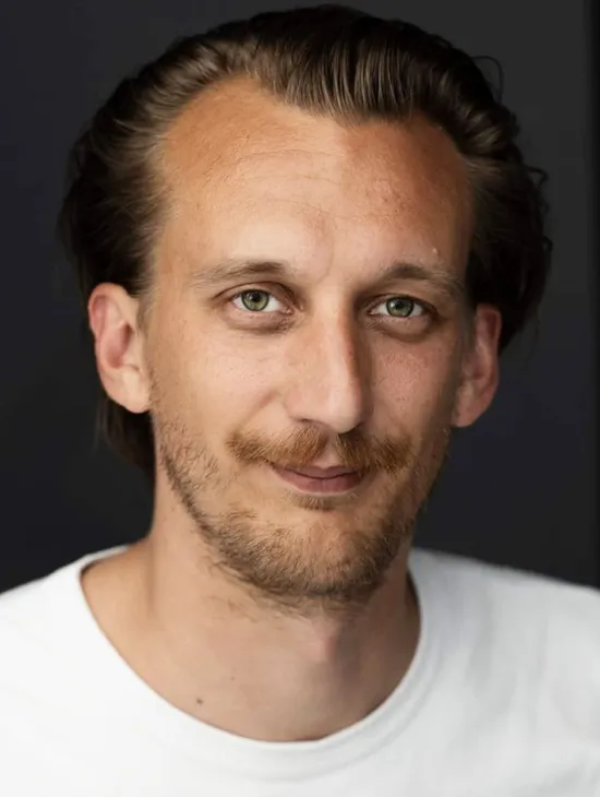 Porträt von Sascha Zauner vor einem dunklen Hintergrund. Er trägt ein weißes T-Shirt, blickt direkt in die Kamera und lächelt leicht.