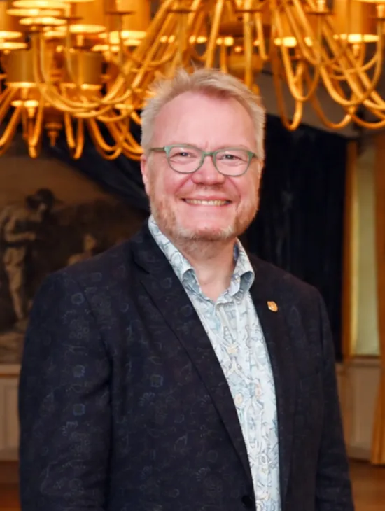 Porträt von Simon Wendring mit Brille, gemustertem Hemd und dunklem Jackett. Er lächelt in die Kamera; im Hintergrund sind goldfarbene Kronleuchter zu sehen.