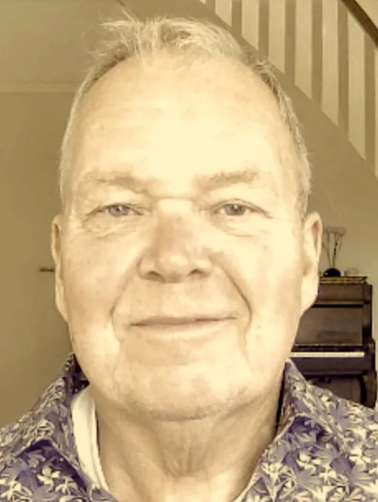 Close-up portrait of Thomas Guthoff wearing a patterned shirt. He smiles slightly at the camera, with a piano and staircase visible in the background.