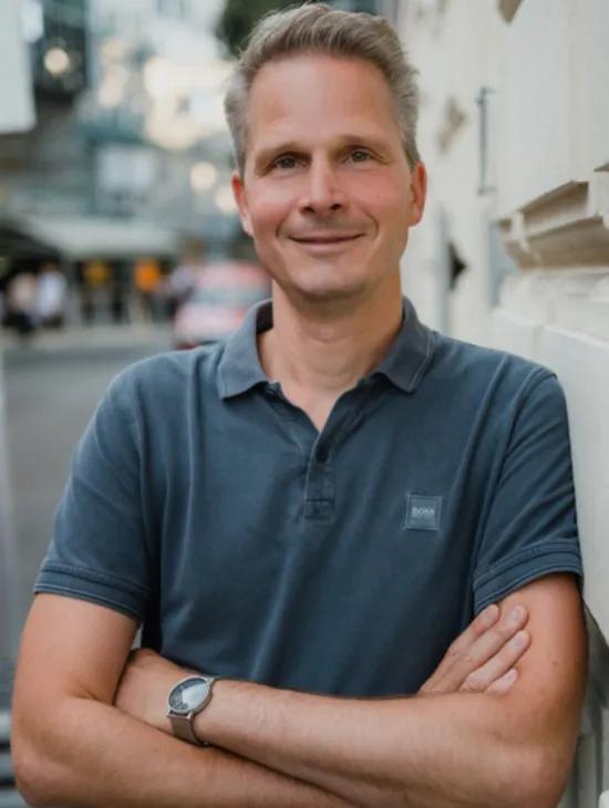 Portrait of Tom Grasshof wearing a dark blue polo shirt. He leans against a building wall with his arms crossed and smiles at the camera.