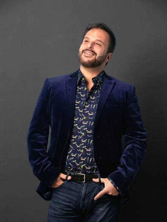 André Heller-Lopes stands against a grey background, smiling and looking upwards. He wears a dark blue blazer over a patterned shirt and has his hands in his trouser pockets.