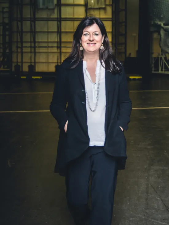 Birgit Meyer steht in einem schwarzen Hosenanzug und einer weißen Bluse auf einer Bühne. Sie hat die Hände in den Taschen und lächelt in die Kamera. Im Hintergrund sind Bühnenaufbauten zu sehen.